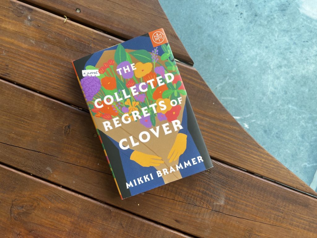 A book titled The Collected Regrets of Clover rests on a stained wooden deck next to a pool.