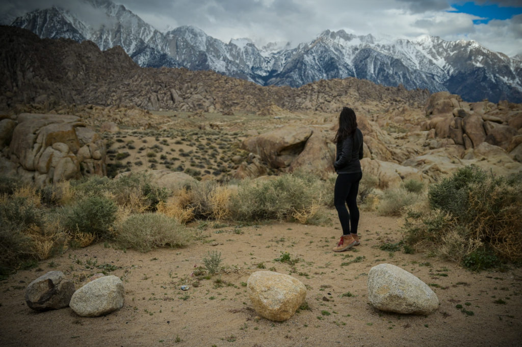 A woman stands with her back toward the camera gazing at snow-capped mountains in the distance.